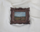 Zoomed out view of the miniature framed landscape painting of a cow grazing in a field, set in an ornate dark bronze frame
