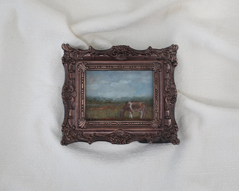 Zoomed out view of the miniature framed landscape painting of a cow grazing in a field, set in an ornate dark bronze frame