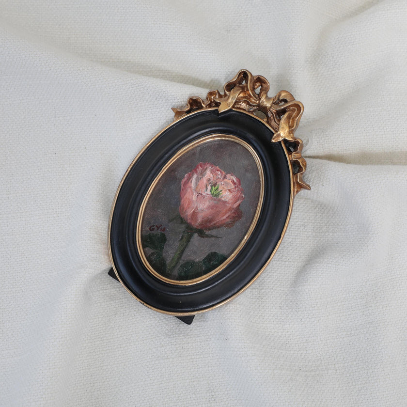 Framed pink rose miniature oil painting resting on cream fabric, showing the gilt bow frame and green leaves
