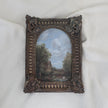 Original antique-style framed landscape painting featuring a rustic cottage, trees, and a cloudy sky, set in an ornate dark brown carved frame on a white fabric background.
