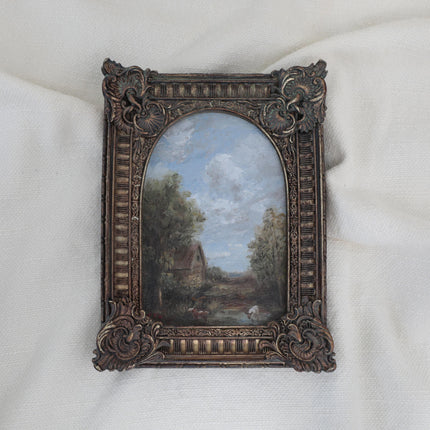 Original antique-style framed landscape painting featuring a rustic cottage, trees, and a cloudy sky, set in an ornate dark brown carved frame on a white fabric background.