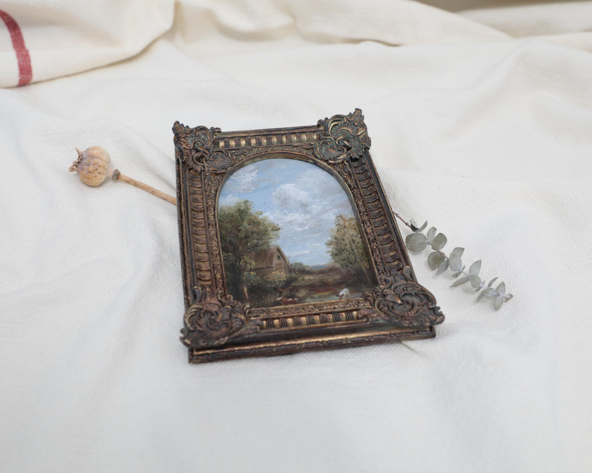 Another angle view of miniature framed landscape painting with an ornate dark brown carved frame, displayed on a draped white fabric background.