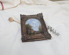 Another angle view of miniature framed landscape painting with an ornate dark brown carved frame, displayed on a draped white fabric background.