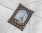 Different angle view of countryside landscape painting of a rustic windmill under a cloudy blue sky in an ornate bronze vintage-style frame
