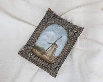 Different angle view of countryside landscape painting of a rustic windmill under a cloudy blue sky in an ornate bronze vintage-style frame