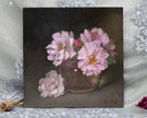 A square oil painting of pale pink flowers in a brown vase against a dark brown background, displayed on a white lace-draped surface with dried botanical props.
