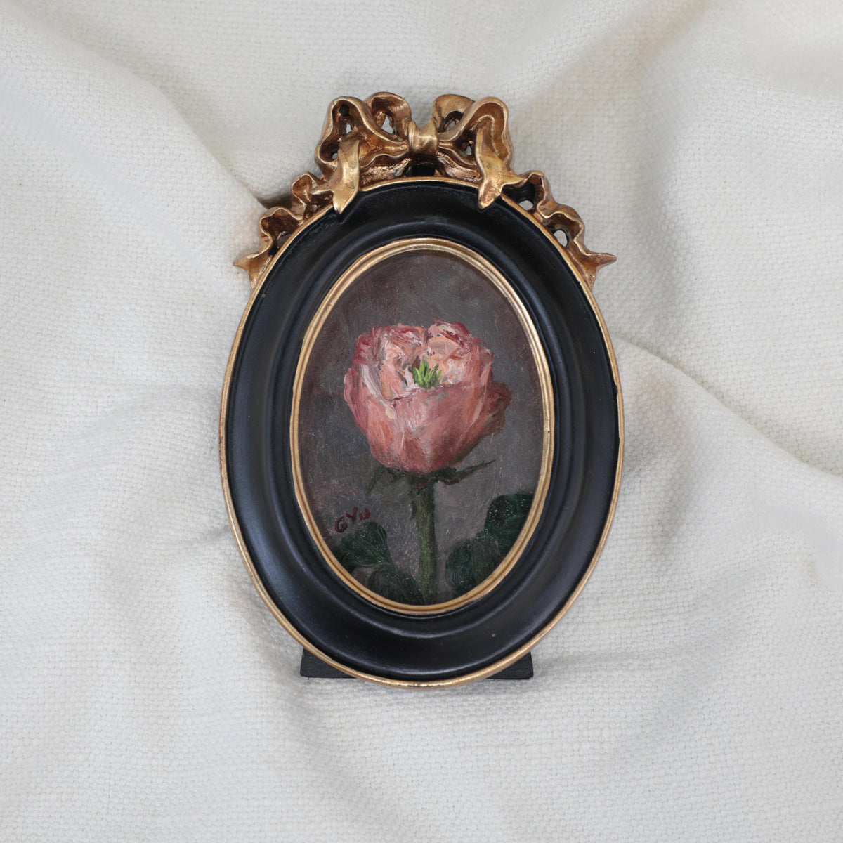 Front view of a miniature pink rose oil painting in a black oval frame with a gilt ribbon bow crest