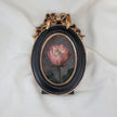 Front view of a miniature pink rose oil painting in a black oval frame with a gilt ribbon bow crest