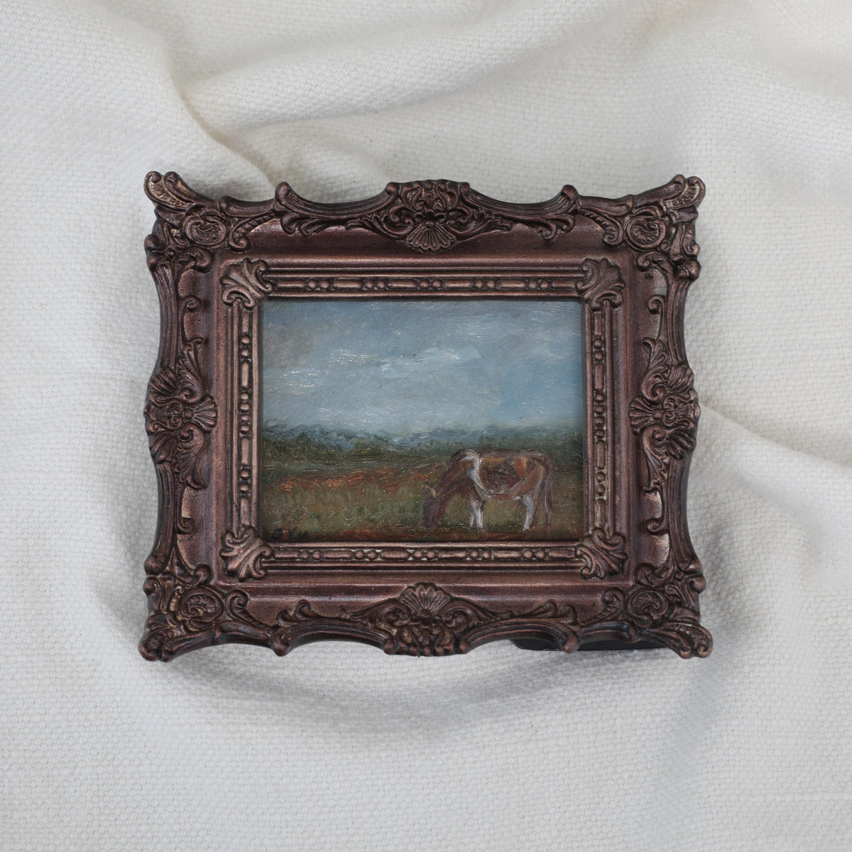 A miniature framed landscape painting of a cow grazing in a field, set in an ornate dark bronze frame and displayed on a white textured fabric background.