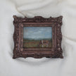 A miniature framed landscape painting of a cow grazing in a field, set in an ornate dark bronze frame and displayed on a white textured fabric background.