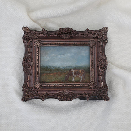 A miniature framed landscape painting of a cow grazing in a field, set in an ornate dark bronze frame and displayed on a white textured fabric background.