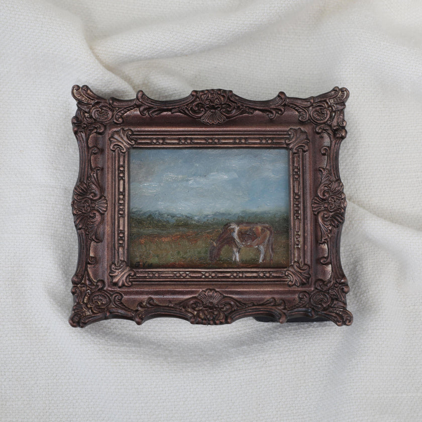 A miniature framed landscape painting of a cow grazing in a field, set in an ornate dark bronze frame and displayed on a white textured fabric background.