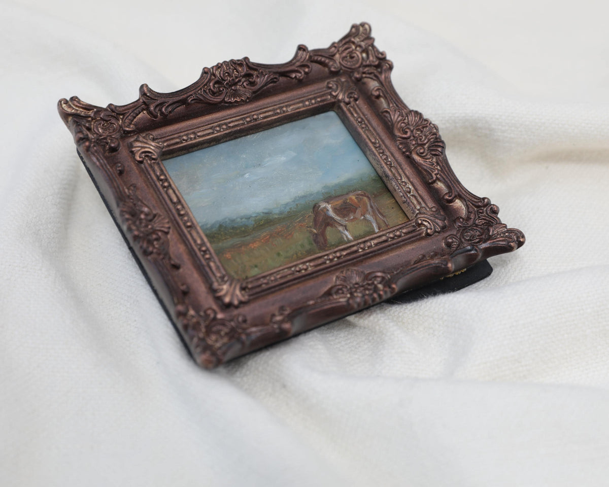 Side view of the miniature framed landscape painting of a cow grazing in a field, set in an ornate dark bronze frame