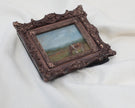 Side view of the miniature framed landscape painting of a cow grazing in a field, set in an ornate dark bronze frame