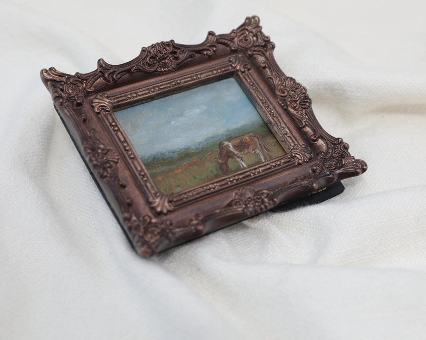 Side view of the miniature framed landscape painting of a cow grazing in a field, set in an ornate dark bronze frame