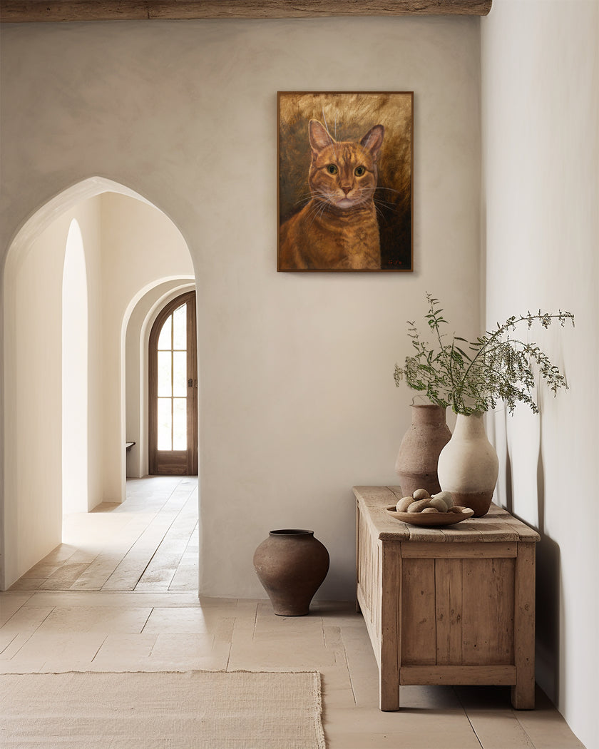A framed portrait painting of an orange tabby cat hangs on a beige plaster wall in a rustic hallway with arches, a wooden console table, and ceramic vases.
