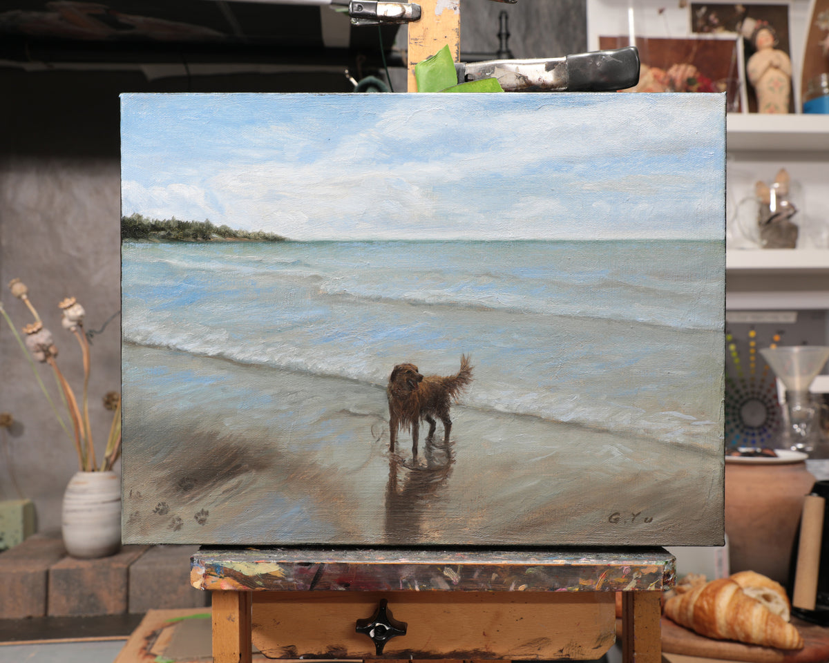A canvas painting of a dog standing on a beach at the water's edge, displayed on an easel in an artist's studio. The painting features a light blue sky, gentle waves, and a sandy beach with the dog's reflection.