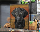 A portrait painting of a black dog on a canvas, displayed on an easel in an artist's studio with paintbrushes and supplies in the background.