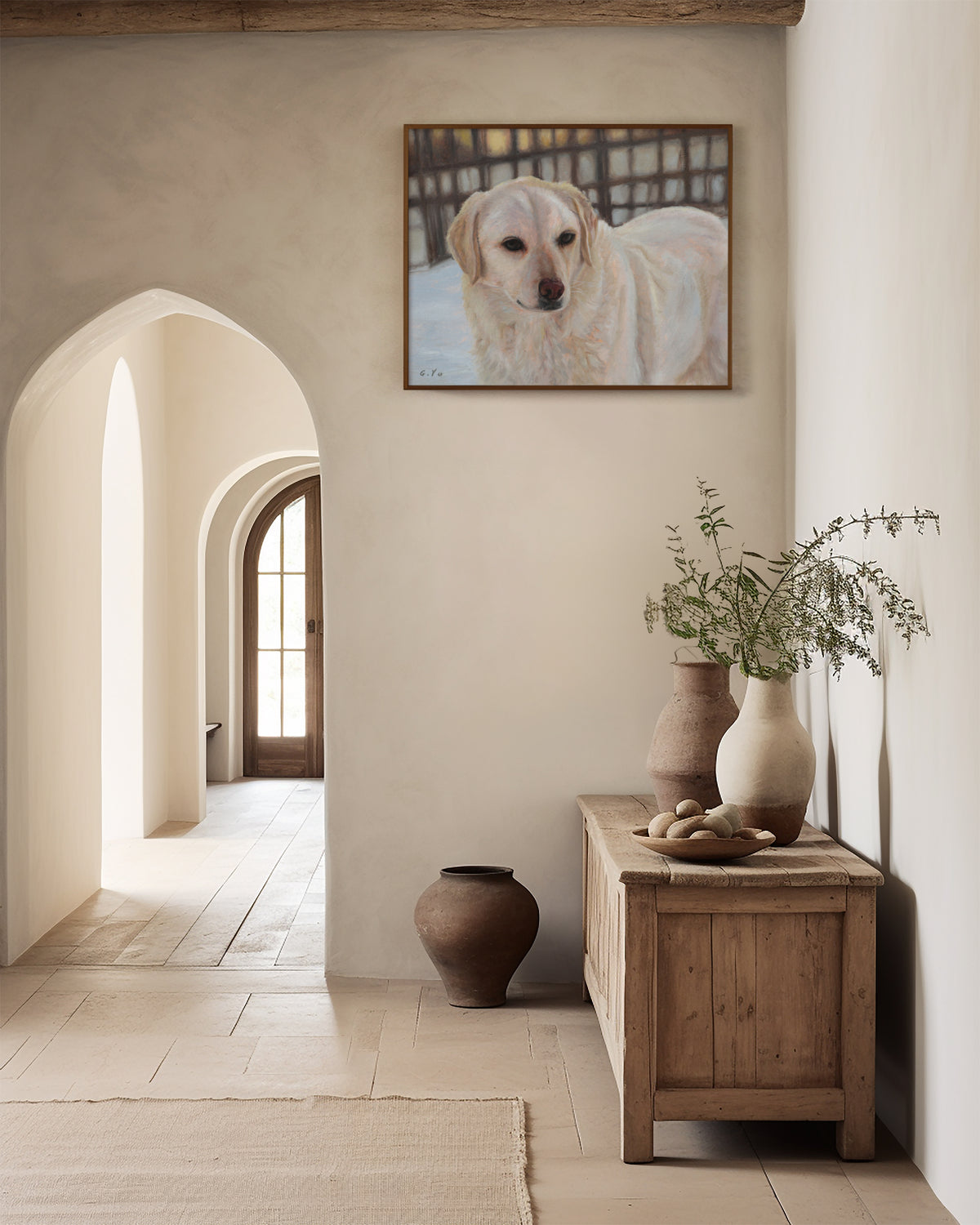 Oil painting of a cream-colored retriever hangs on a warm beige plaster wall in a rustic, sunlit room with arched doorways, a wooden console table, and ceramic vases.