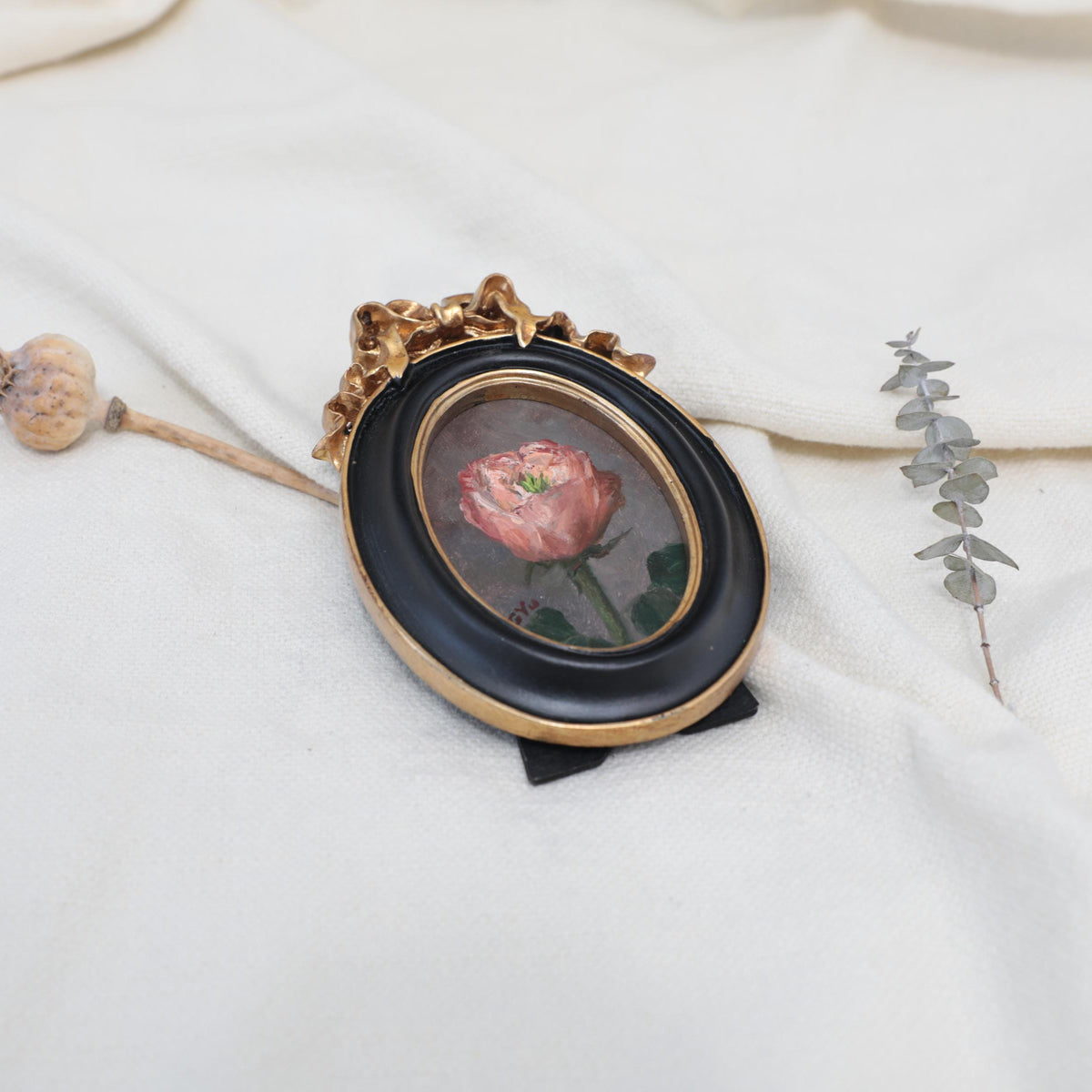 Side view of the original pink rose oil painting in an ornate oval gilt frame on cream linen with a dried poppy pod and eucalyptus
