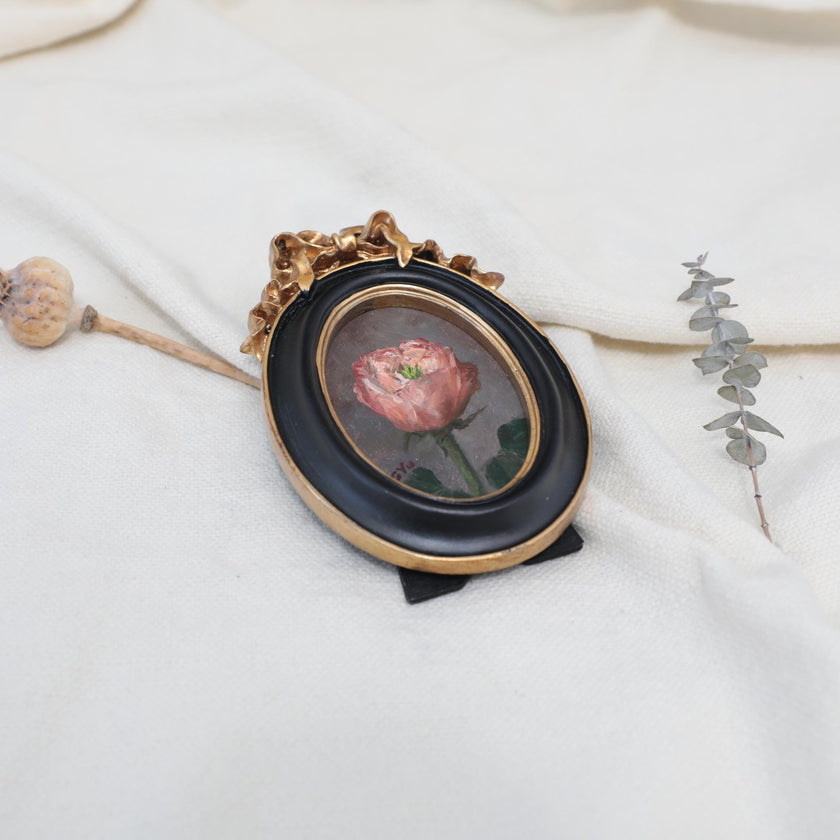 Side view of the original pink rose oil painting in an ornate oval gilt frame on cream linen with a dried poppy pod and eucalyptus