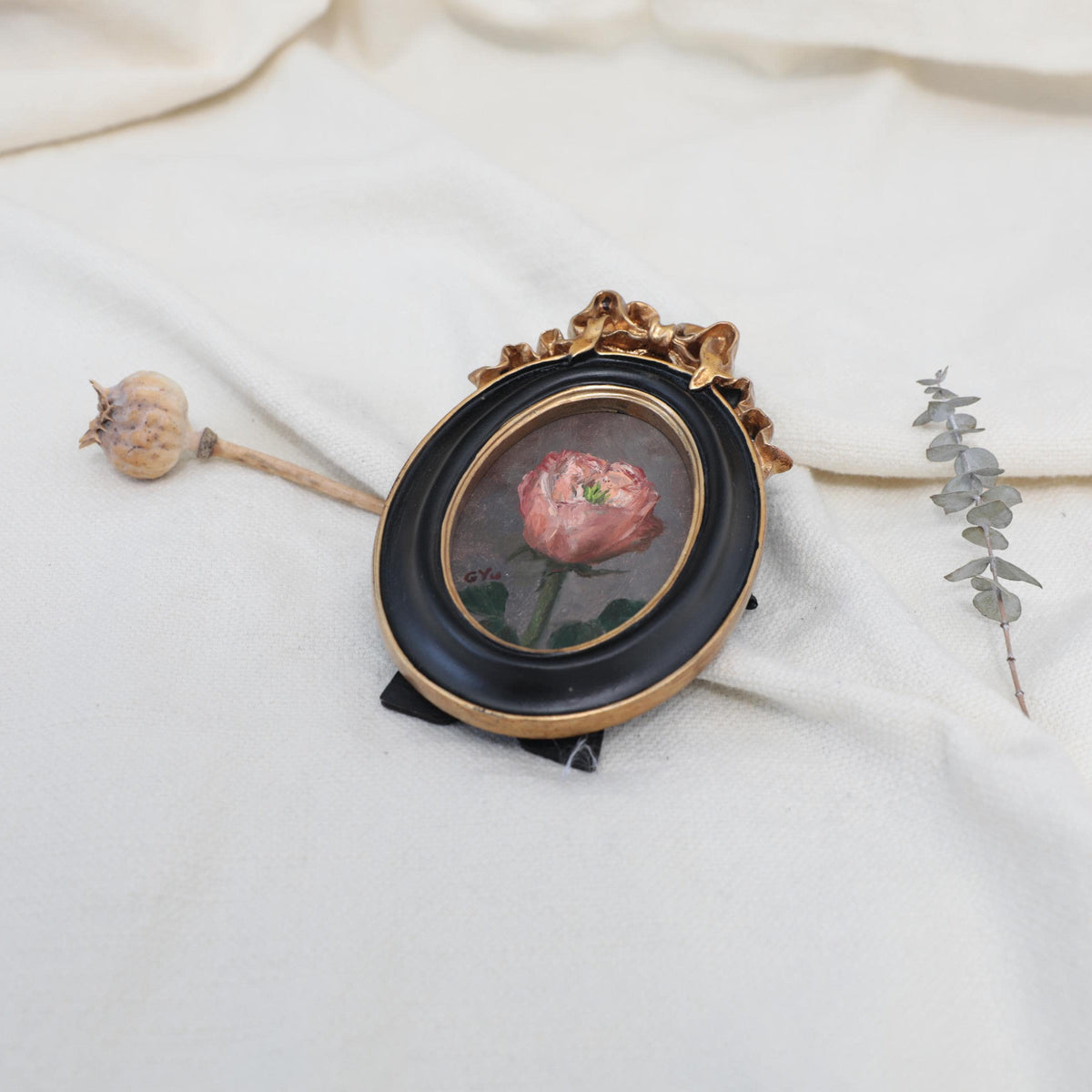 Original pink rose oil painting in an ornate oval gilt frame on cream linen with a dried poppy pod and eucalyptus
