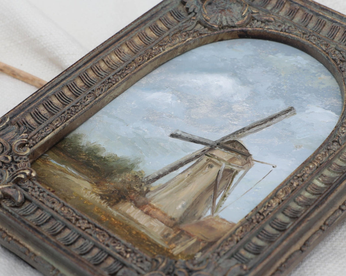 Close up of the countryside landscape painting of a rustic windmill under a cloudy blue sky in an ornate bronze vintage-style frame