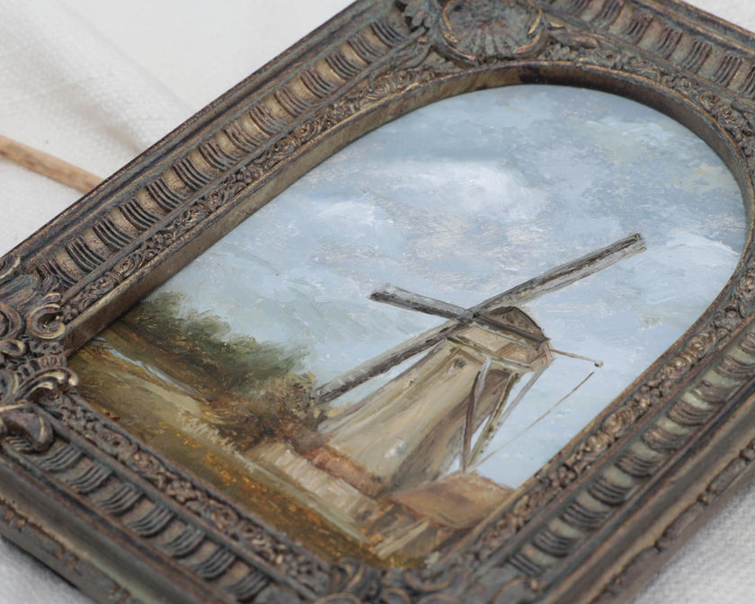 Close up of the countryside landscape painting of a rustic windmill under a cloudy blue sky in an ornate bronze vintage-style frame