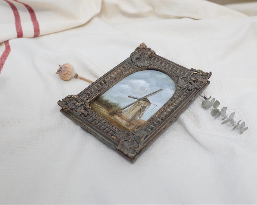Side view of Original countryside landscape painting of a rustic windmill under a cloudy blue sky in an ornate bronze vintage-style frame, with dried flowers for scale. 