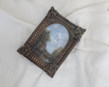 Miniature antique-style framed landscape painting with an ornate dark brown carved frame, displayed on a draped white fabric background.