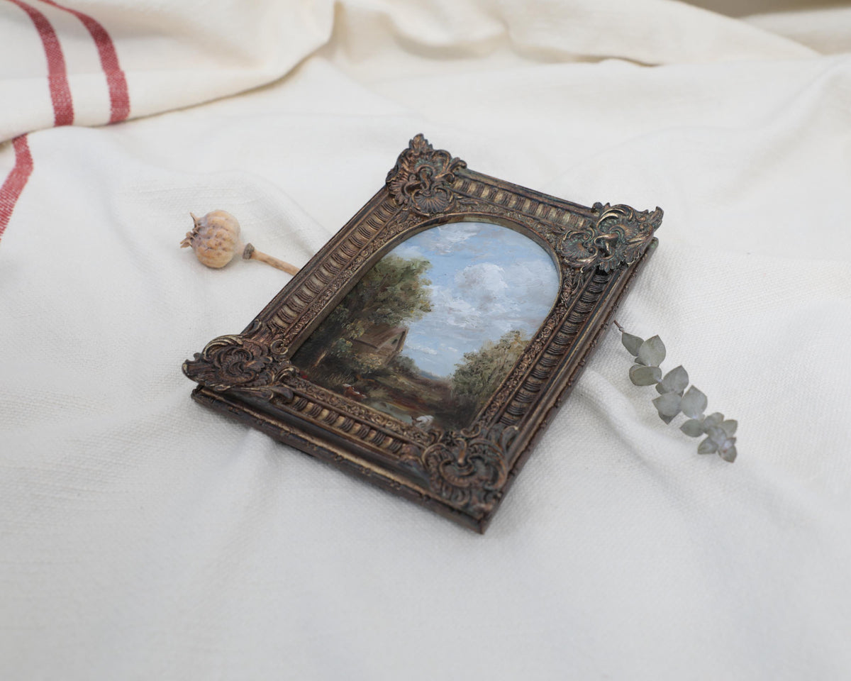 Side view of the original miniature antique-style framed landscape painting with an ornate dark brown carved frame, displayed on a draped white fabric background.