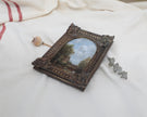 Side view of the original miniature antique-style framed landscape painting with an ornate dark brown carved frame, displayed on a draped white fabric background.