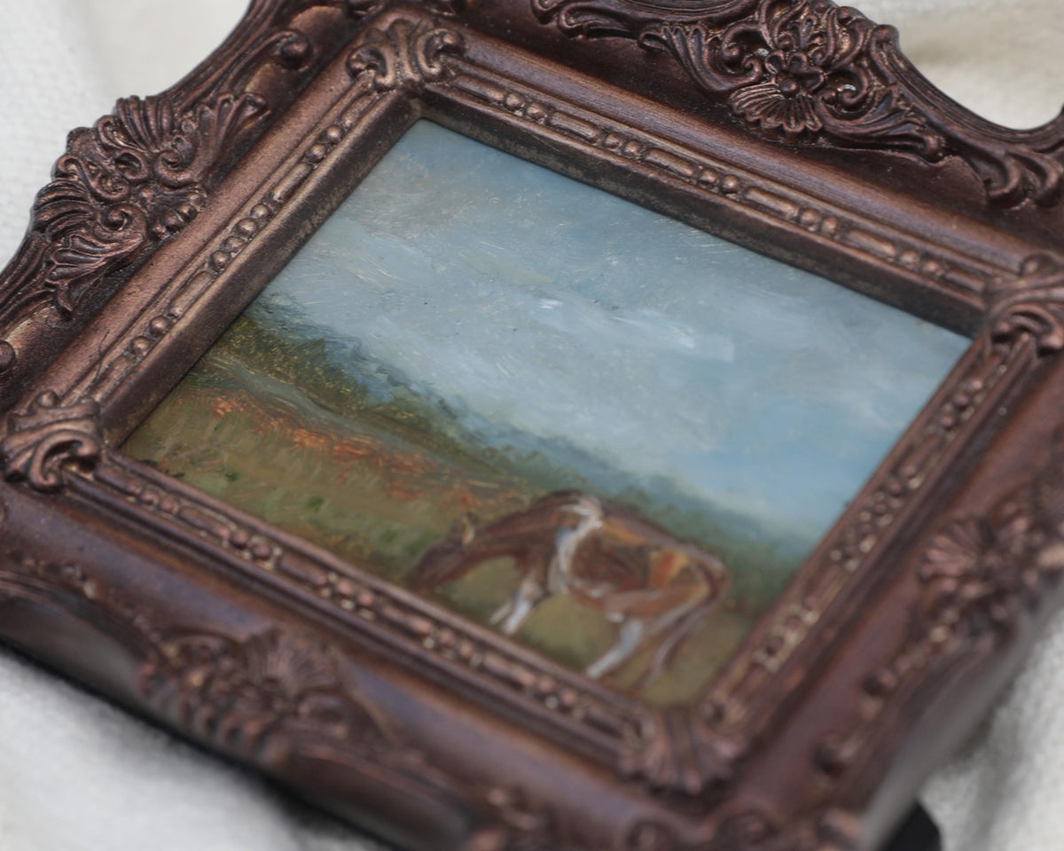 Close up from the side view, of the miniature framed landscape painting of a cow grazing in a field, set in an ornate dark bronze frame