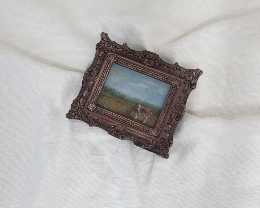 Different angle of the miniature framed landscape painting of a cow grazing in a field, set in an ornate dark bronze frame