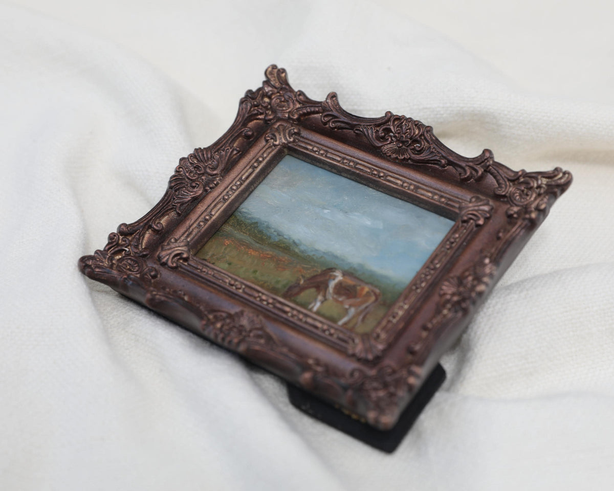 Different side view of the miniature framed landscape painting of a cow grazing in a field, set in an ornate dark bronze frame