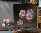 The oil painting of pink flowers in a glass vase, displayed on a wooden artist easel, with the live flower setup at the background. 