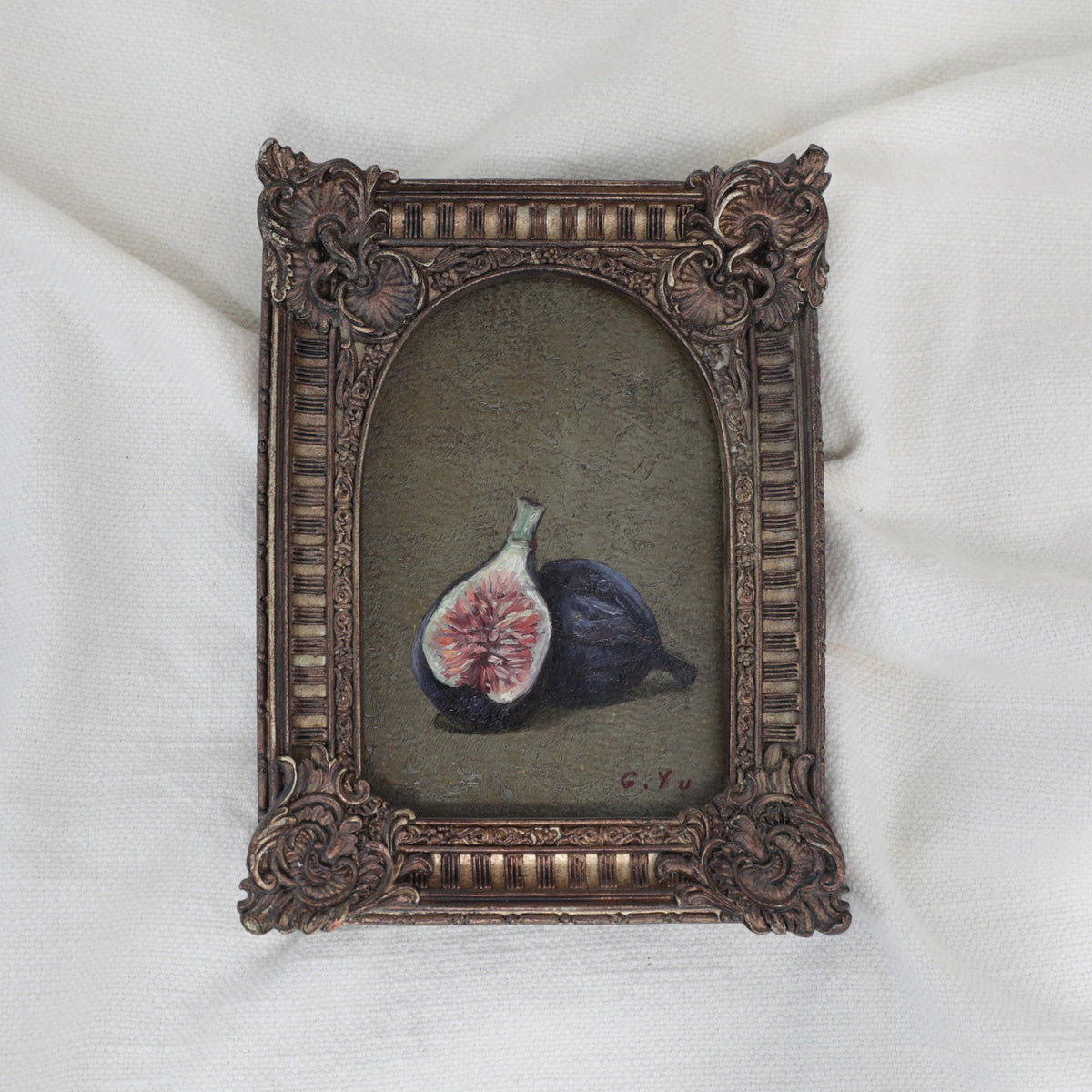 Small still life oil painting of two purple figs, one halved, on an olive-green background in an arched ornate vintage-style frame