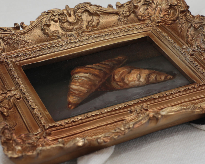 Two croissants oil painting in an antique gold frame, laid flat with soft natural shadow
