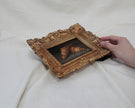 Hand holding the ornate gold frame of a croissant oil painting on a cream fabric backdrop