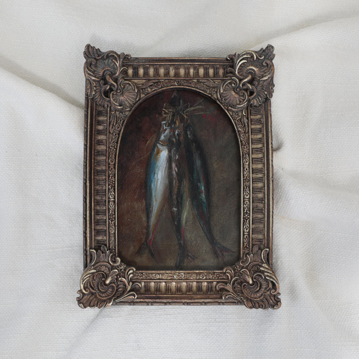 Small still life oil painting of three hanging fish on a dark background in an arched ornate vintage-style frame