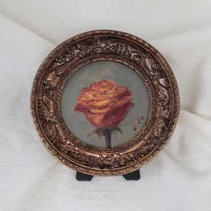 Miniature oil painting of an orange-red rose on a gray-green background in a round ornate bronze frame with grapevine details