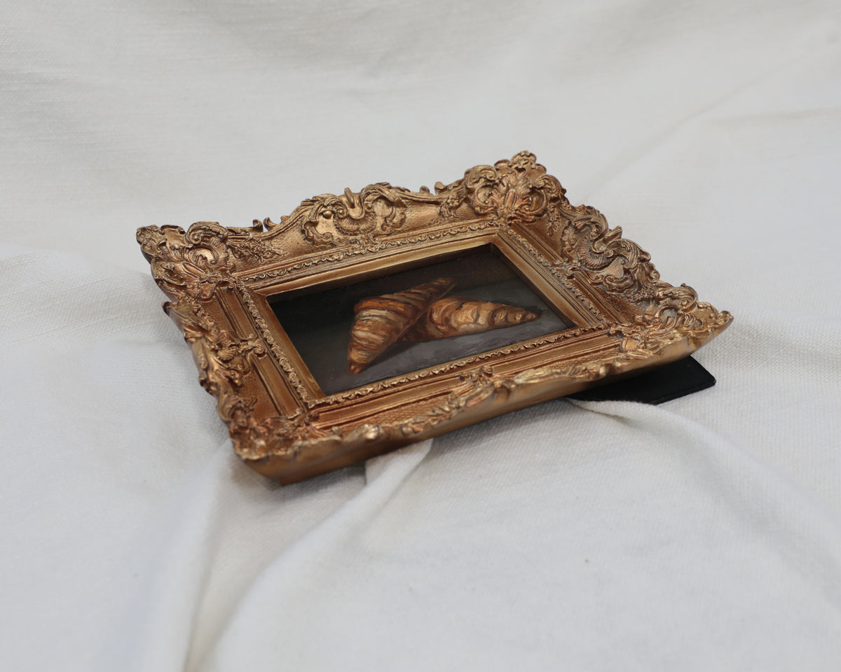 Croissant still life oil painting propped at an angle, showing the frame's fold-out stand