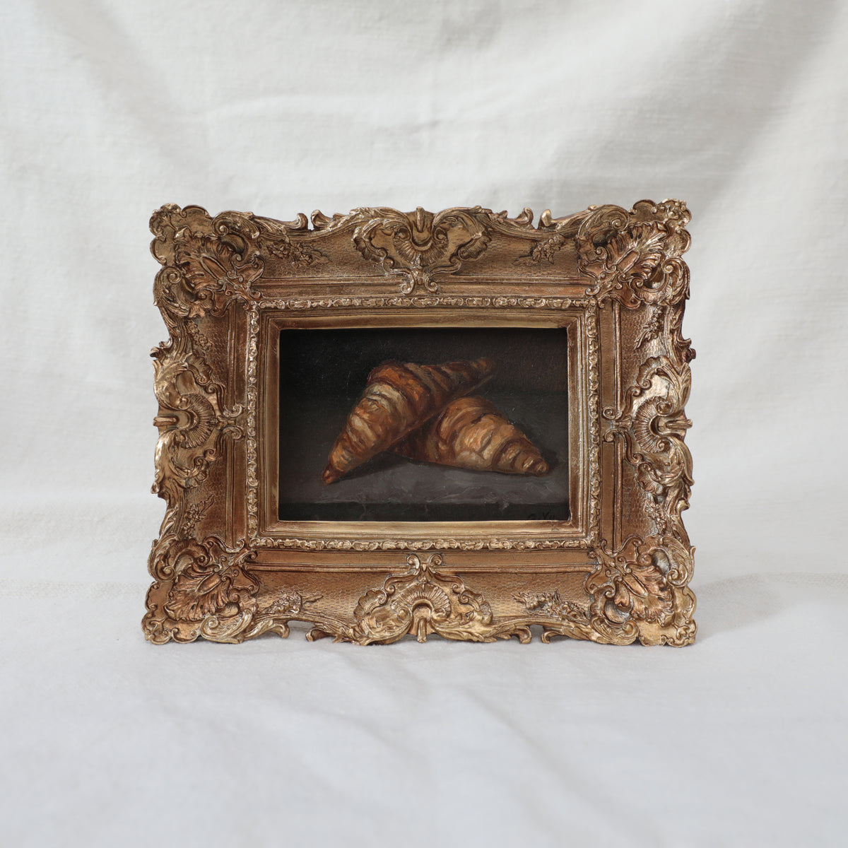 Original oil painting of two croissants on a stone ledge, in an ornate antique gold frame, shown at an angle on a cream fabric backdrop