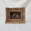 Original oil painting of two croissants on a stone ledge, in an ornate antique gold frame, shown at an angle on a cream fabric backdrop
