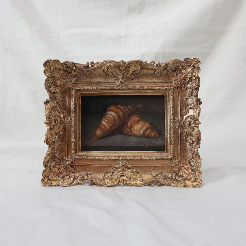 Original oil painting of two croissants on a stone ledge, in an ornate antique gold frame, shown at an angle on a cream fabric backdrop