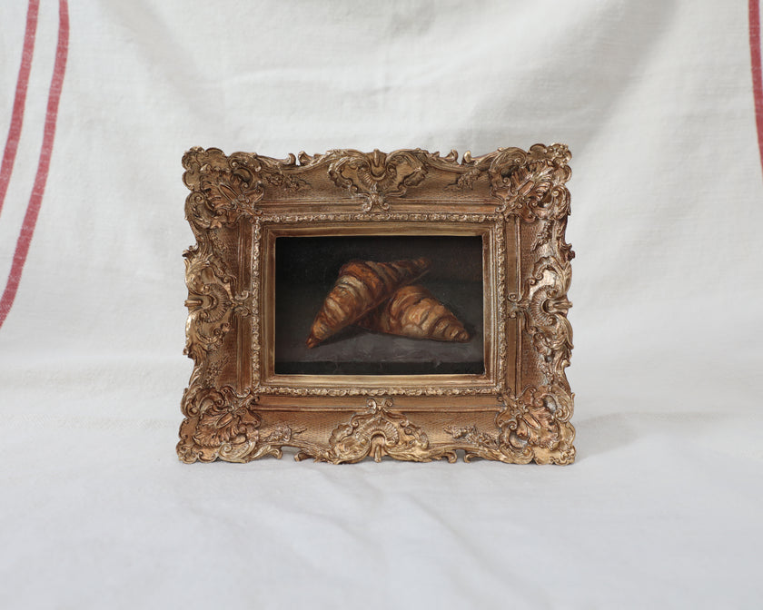 Framed croissant oil painting shown beside striped linen, with the full ornate gold frame visible