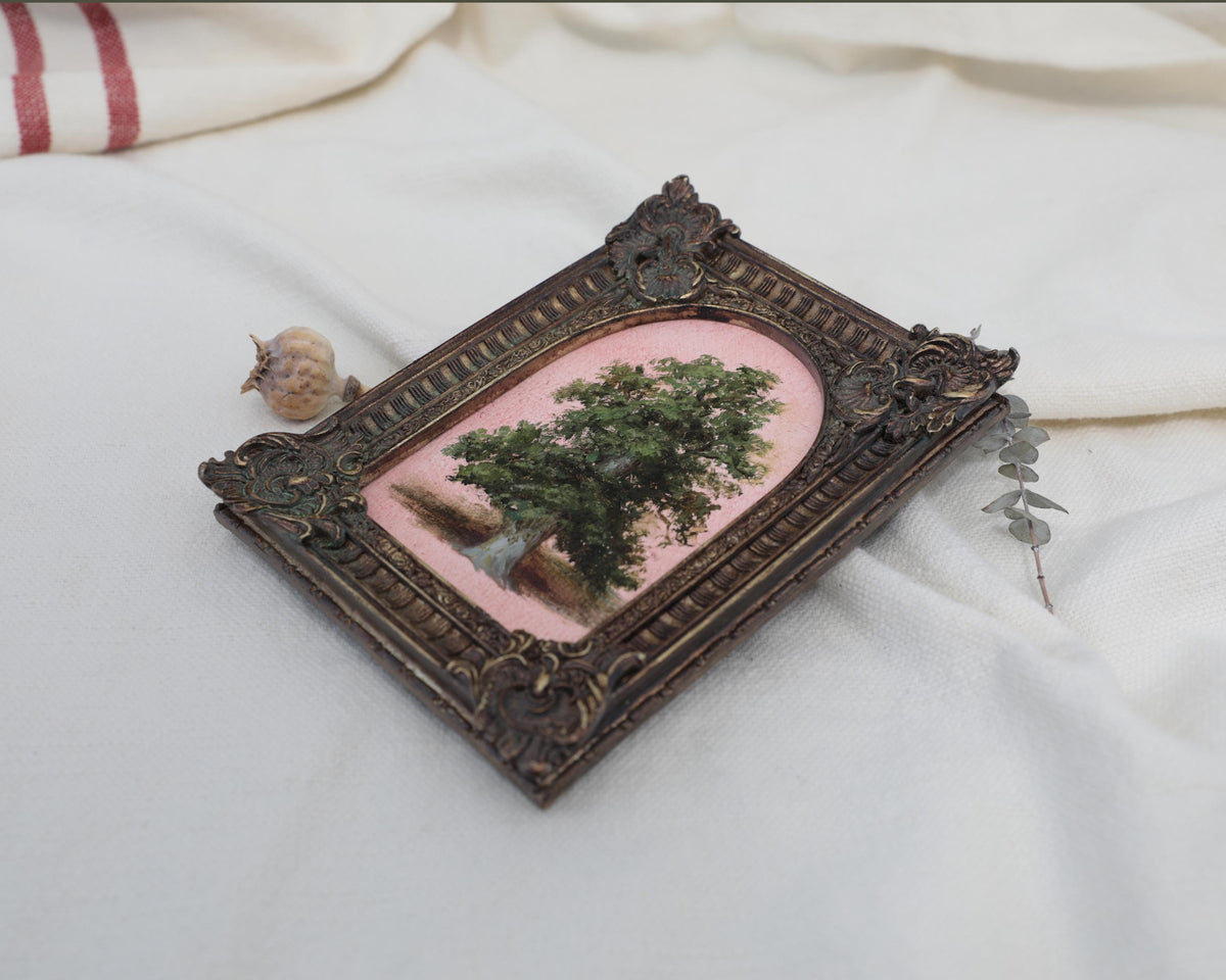 Angled view of a vintage-style tree oil painting in an ornate frame beside dried flowers